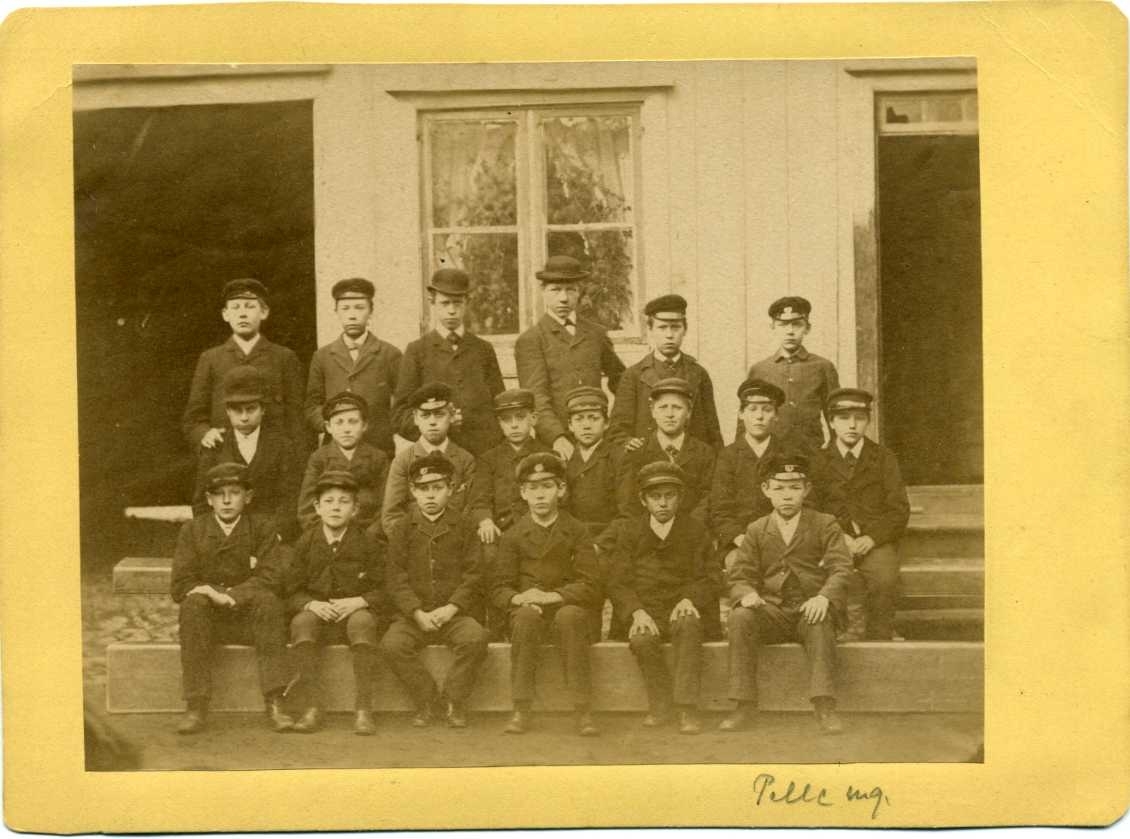 En skolklass med pojkar, två sittande led och ett stående - de äldre. Alla bär keps eller hatt - de äldre.