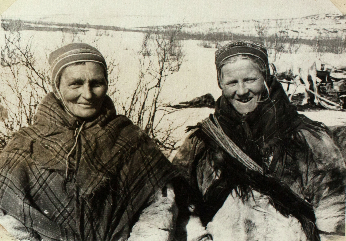 Bildet viser to samiske kvinner, ikledd pelskofter, sjal og gákti. En av dem har en lasso rundt. Begge ser inn i kameraet og smiler bredt. I bakgrunnen ser man reinsdyr med sleder og trær og busker i snødekt landskap.