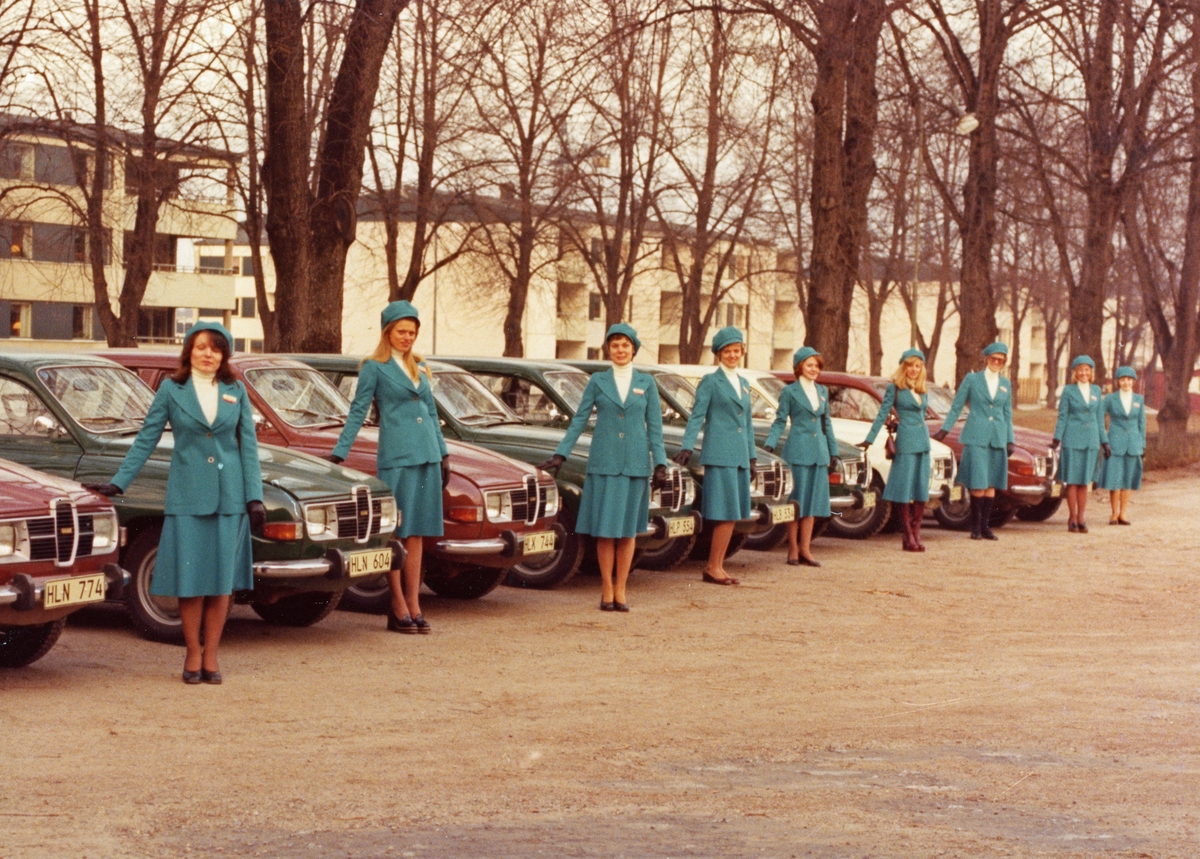 Bilpatrullen. 9 kvinnor i blå dräkter med kjol, kavaj samt keps framför varsin SAAB. Stadsdelen Öster i bakgrunden.