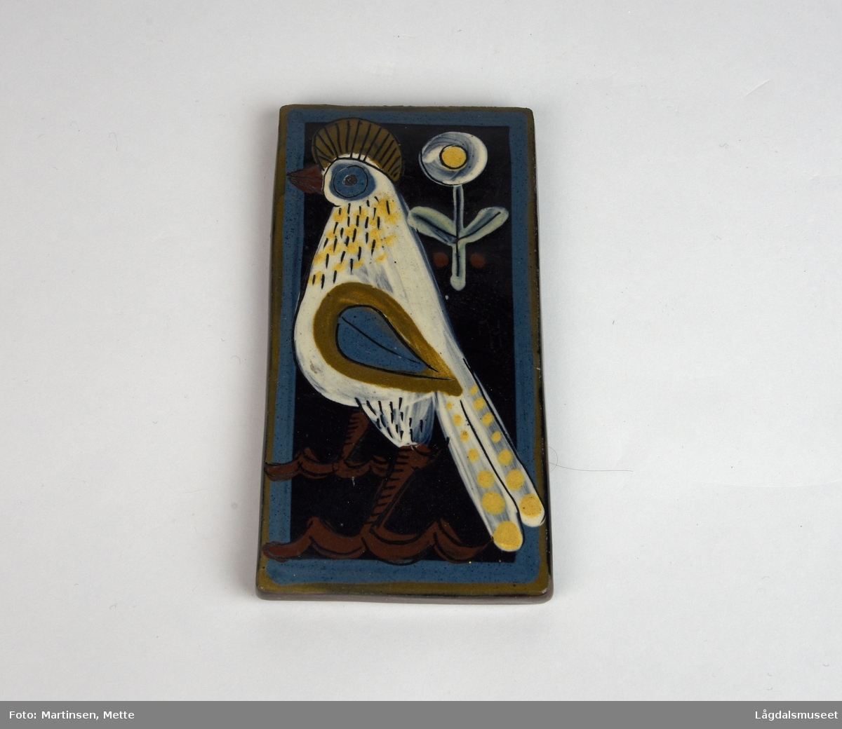 Rektangulær keramikkflis. Motivet er en hvit fugl og hvit blomst. Mørk blå bakgrunn og innrammet av en lysere blå kant.