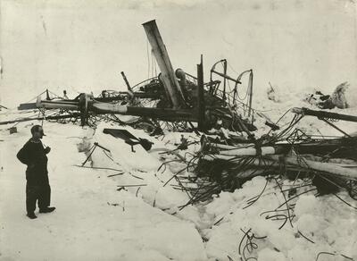 Sorthvitt bilde med en mann til venstre og et skipsvrak i isen til høyre. Det er mye snø på bildet