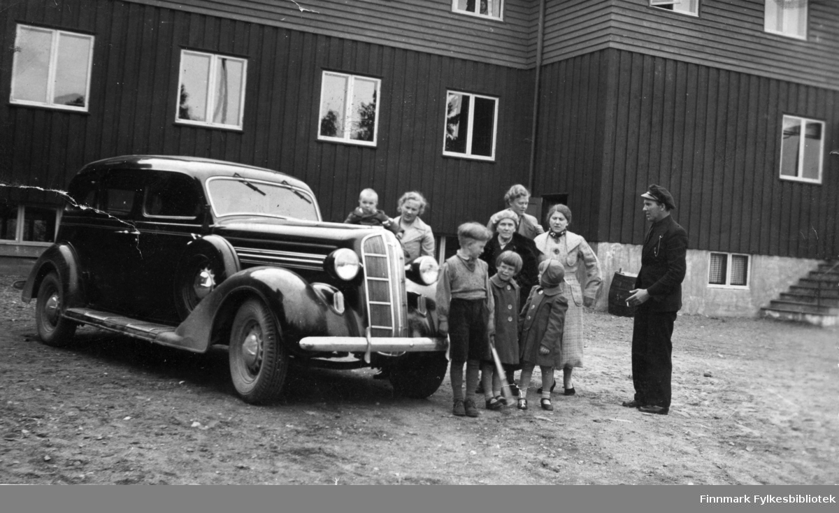 Utenfor Svanvik Folkehøgskole. Flere personer, både voksne og barn står ved siden av en Dodge 1936 modell. Foran står fra venstre: Martin Seljemo, tvillingene Ruth Seljemo, og Inger Seljemo.