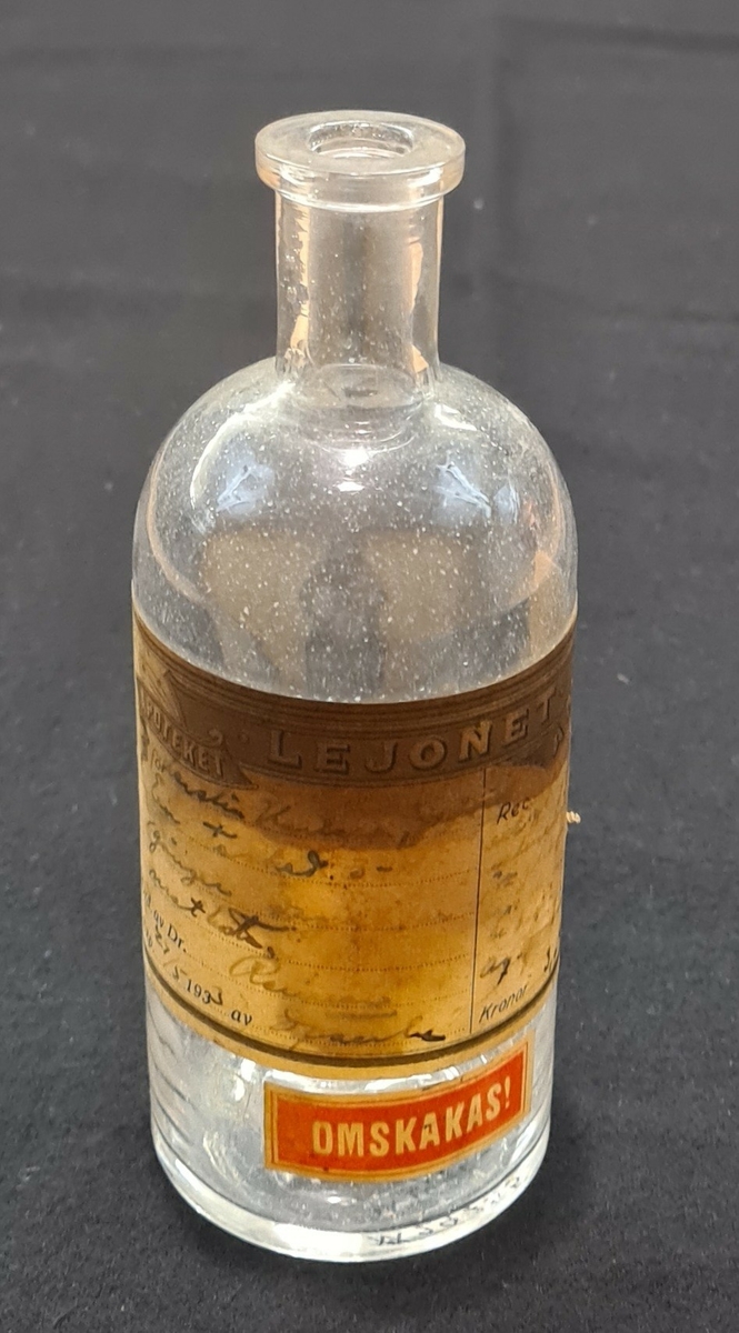 Glasflaska. Vit etikett med handskriven text. Flaskan kommer från Apotek Lejonet i Åmål. Ytterligare en etikett finns på flaskan där det står "OMSKAKAS!"

I botten på flaskan står L 200.