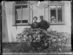 Kvinne og mann stående foran bolighus. I vinduet til høyre s