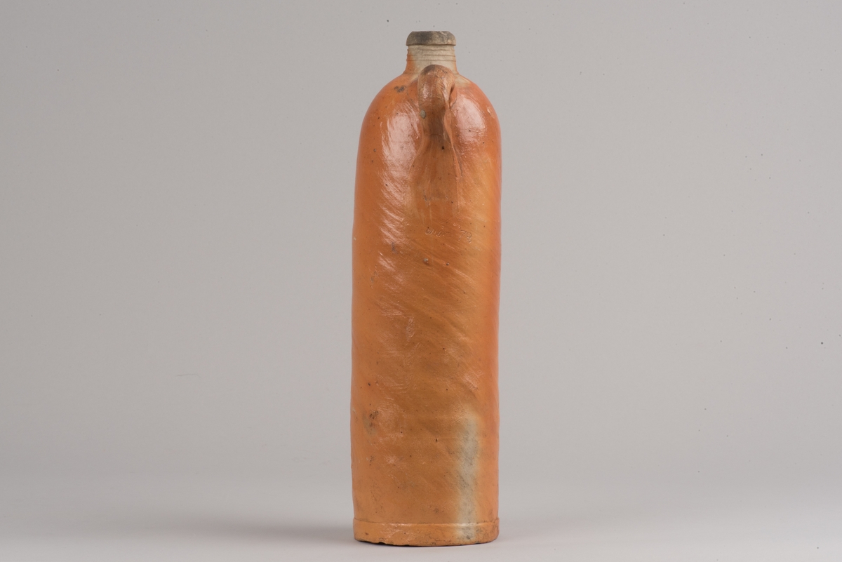 Krus i form av flaska tillverkat av saltglaserat stengods.
Cylindrisk flaska med kort hals och handtag. På buken finns två stämplar som visar att det innehållit seltersvatten.