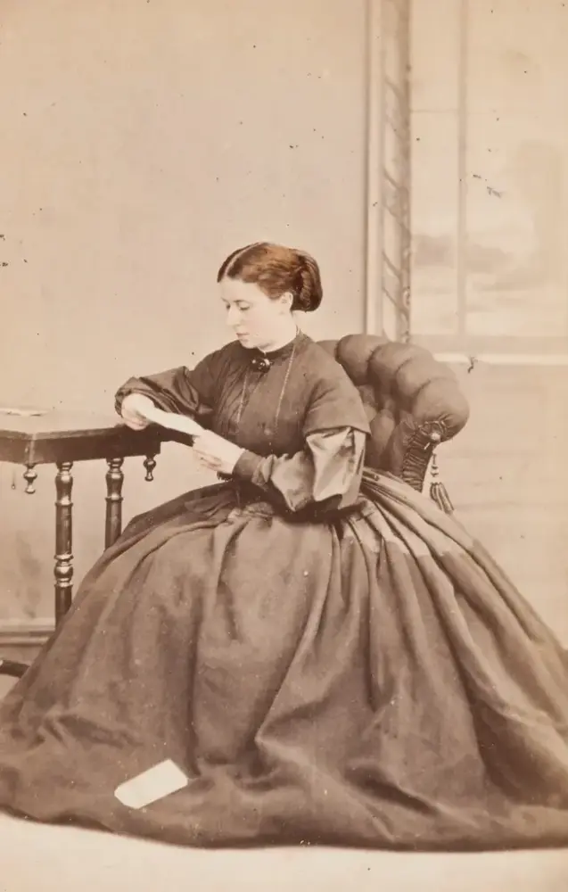 A woman in a large dress sits at a table and reads a letter.