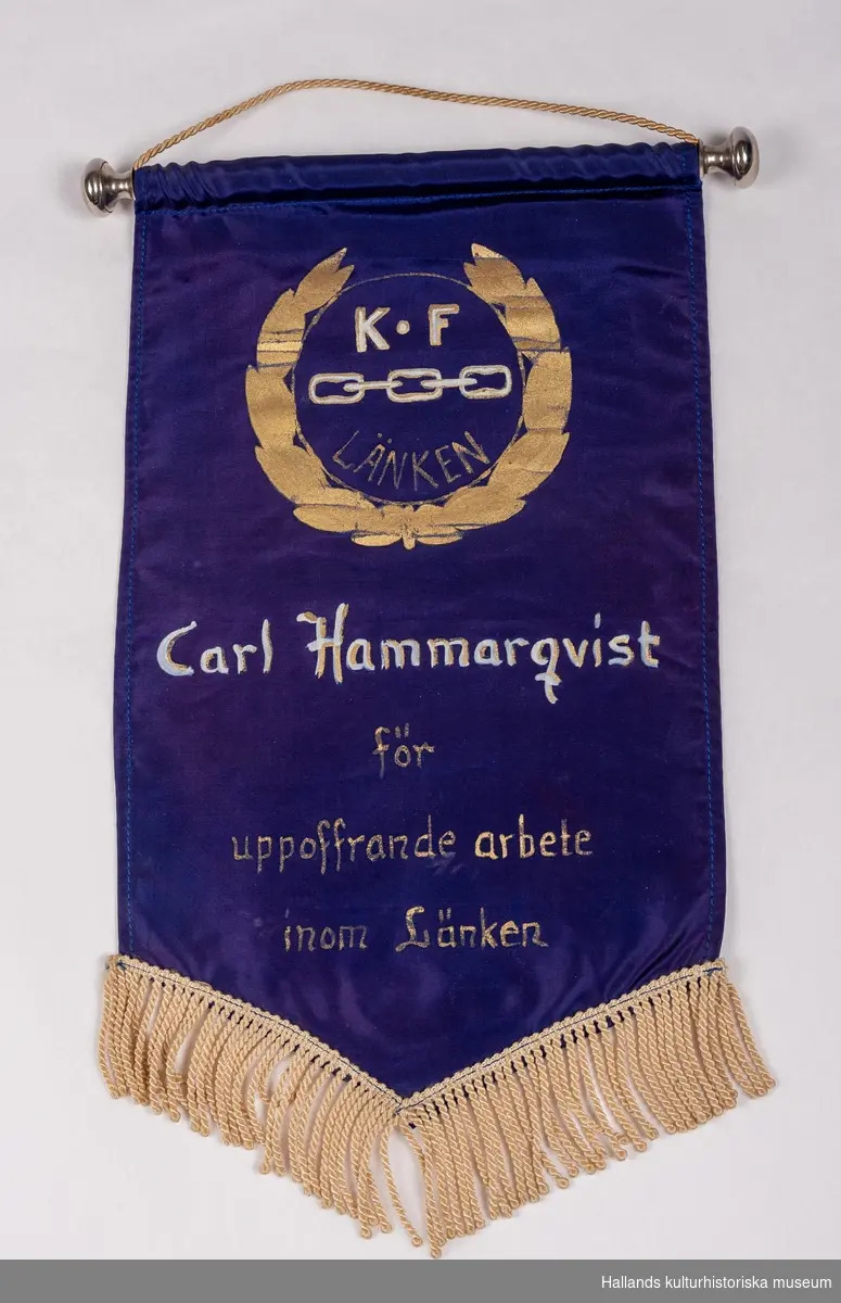 Entungat standar i siden med målad text och emblem "K.F-LÄNKEN, Carl Hammarqvist för uppoffrande arbete inom länken". Silkesfrans nedtill, montering med tunnel och stång med knoppar.