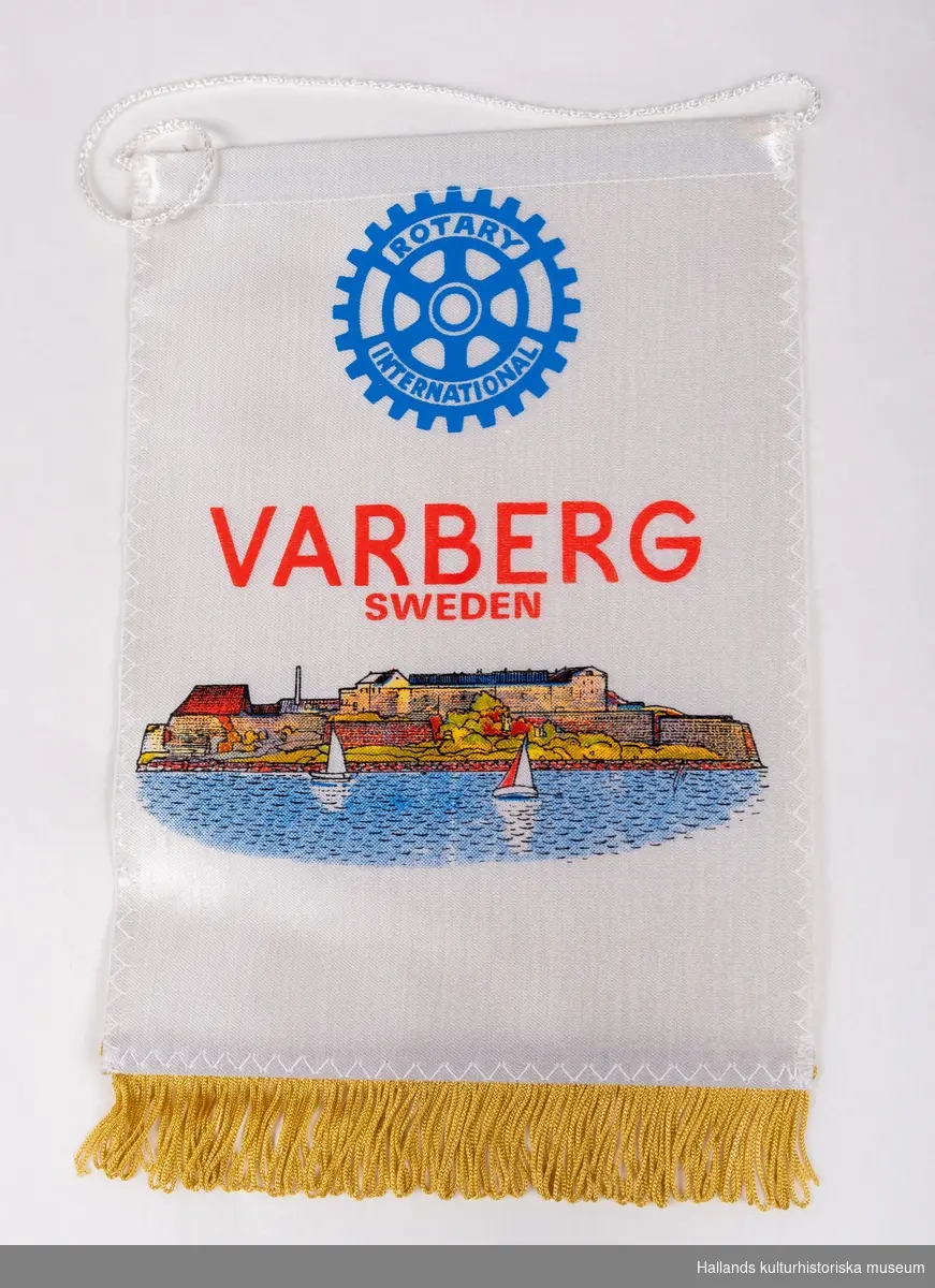Vitt dubbelt silkestyg med påtryckt Rotarys märke och en bild i färg av Varbergs fästning. Text: "Rotary International, Varberg, Sweden.". Fransar nedtill, tråd för upphängning upptill."