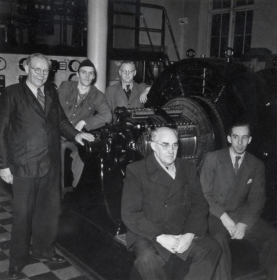 Gruppfoto av några män framför en generator (?) på Växjö elverk, 1949.
Främst sitter maskinmästare Johan Åström och Narin.
Bakom dem fr. v.: E Johansson, Andersson, Bard (enligt text under fotot).