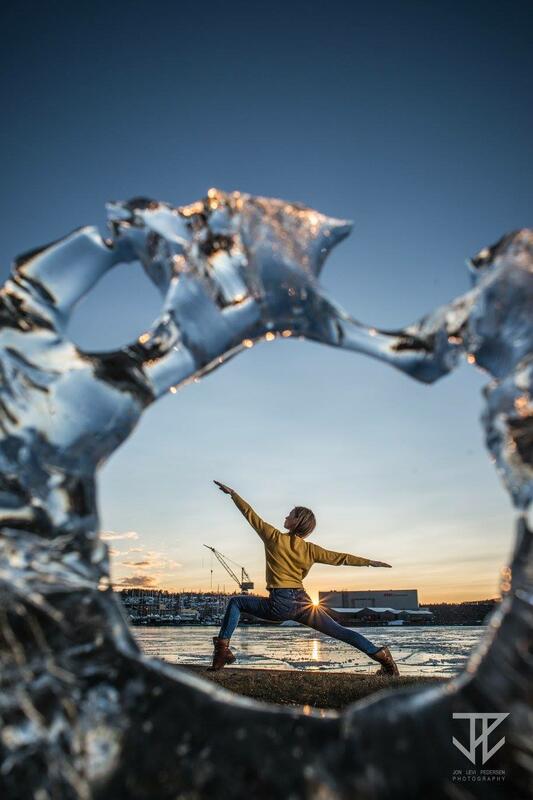 En kvinne i yogaposituren kriger to, fotografert i vinterlandskap i solnedgang gjennom en isskulptur som utgjør en sirkel.