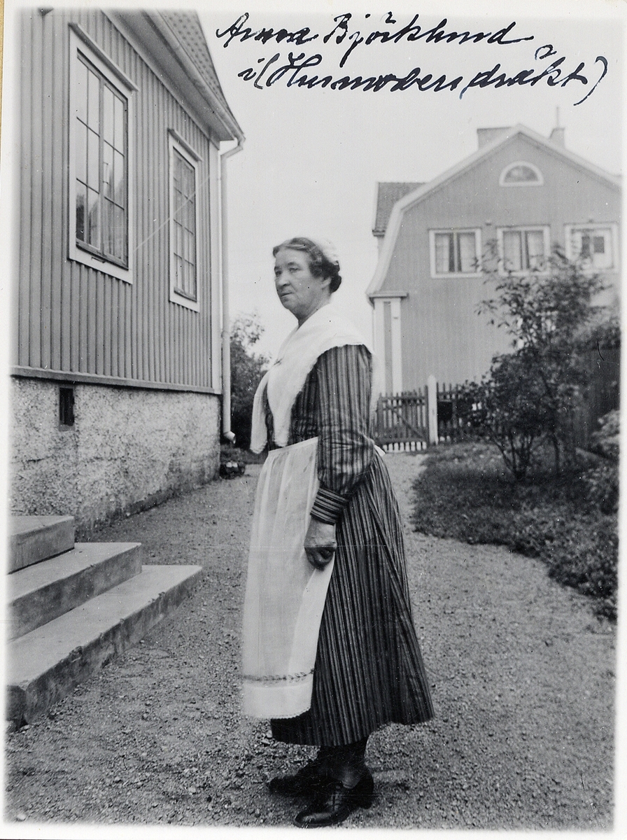 En kvinna står utanför ett bostadshus, iförd s.k. husmorsdräkt.