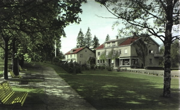 Kolorerat vykort, "Torgparken, Hyltebruk". Längs den stenlagda gången i parken står bänkar och vid gatan ligger tvåvåningshus med bostäder och butiker.