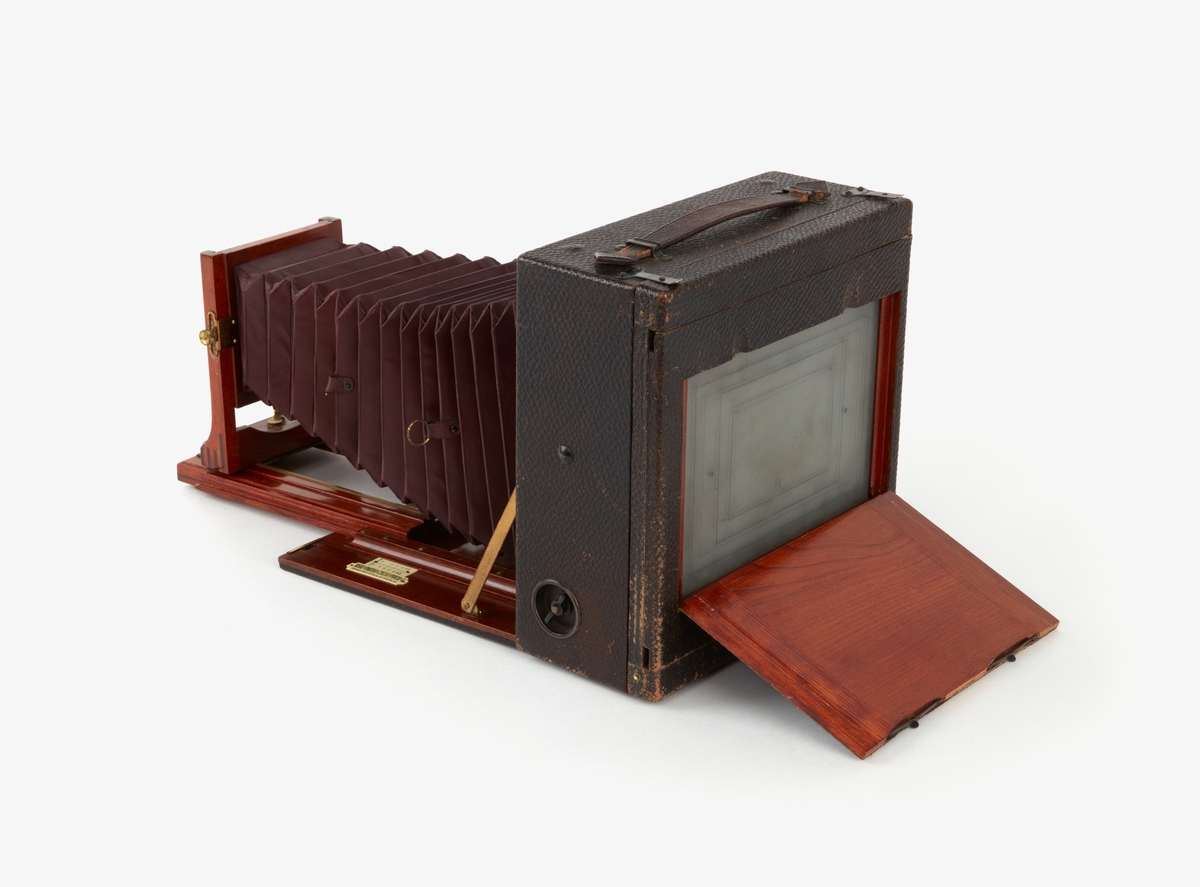 Portabel bälgkamera som förvaras i en svart låda som ligger i väska av ljust skinn. Text: TELE-PHOTO POCO. Väskan saknar axelrem.