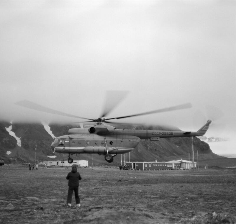 Helicopter during landing. One person stands in front, with the back towards the camera. Mountains in the background.