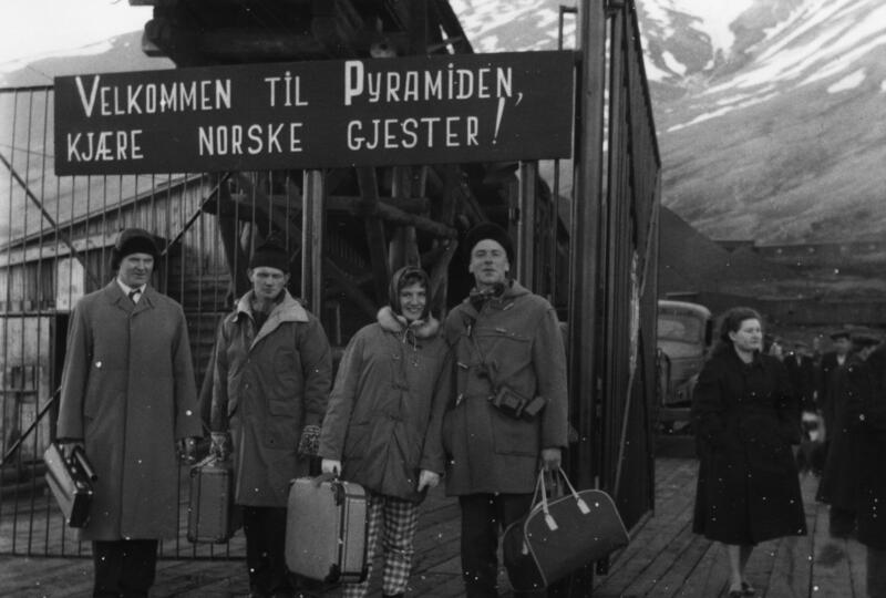 Bilde av fire personer i vinterklær foran et skilt der det står Velkommen til Pyramiden, kjære norske gjester