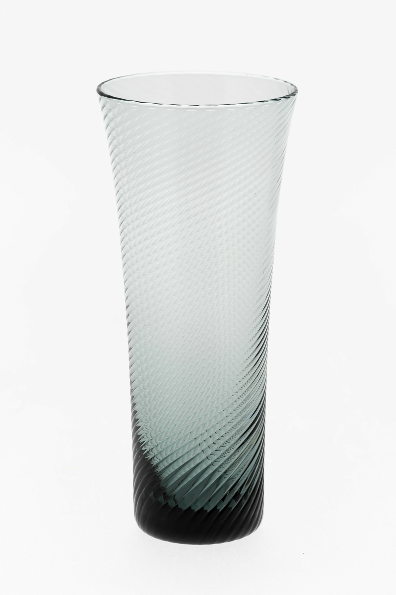 Seltersglass i gråfarget gjennomskinnelig glass. Svak konisk form med sirkulær munningsrand, hvor spiralformede riller snor seg langs korpus.