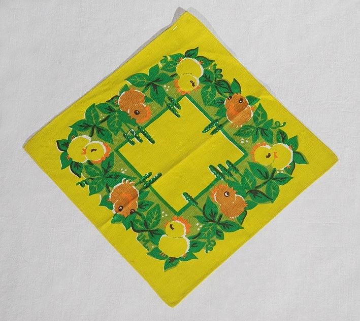 Citrongul, kvadratisk påskduk med tryckt mönster av citrongula repektive bruna kycklingar, samt gröna bladslingor med fröhängen i en kvadrat mellan dukens ytterkant och mittparti.