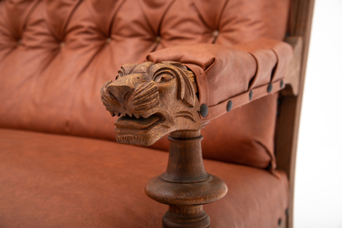 Sofa med armlener med utskåret "løve"-hode på enden av armlenet. 2 dreide forbein . Trukket med imitert skinn; skai. Polstring i chesterfield-stil med dyphefting.