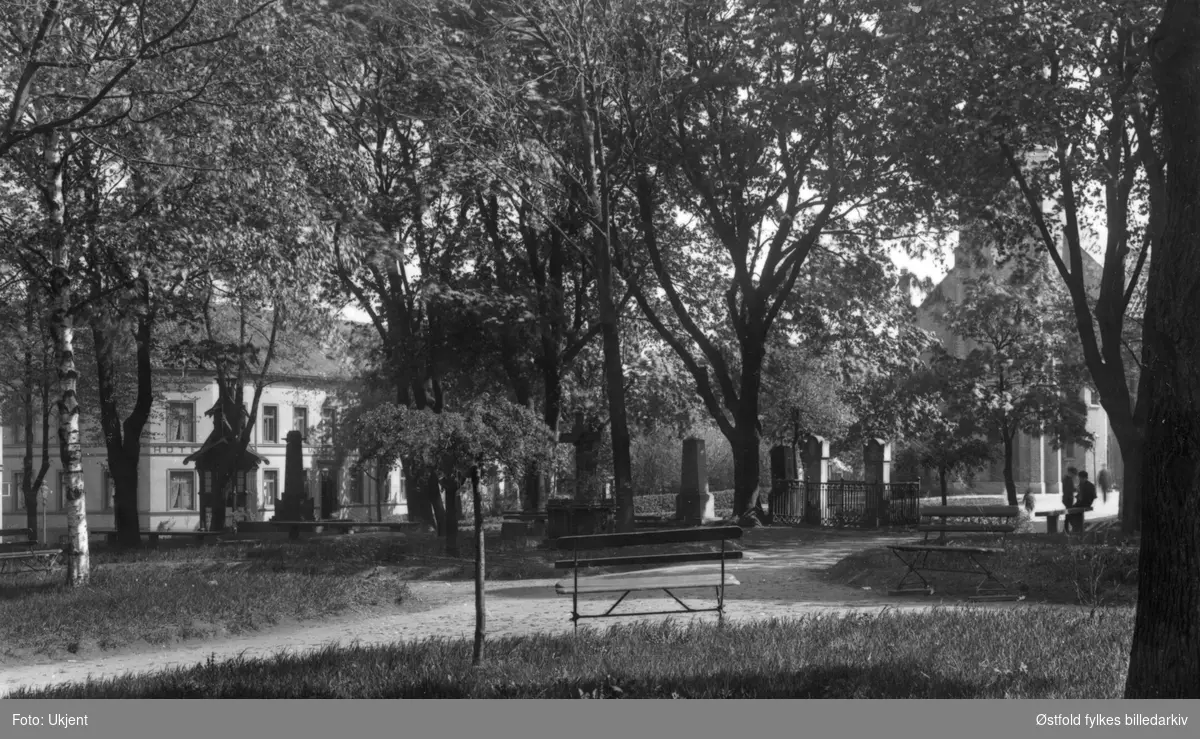 Kirkeparken i Moss ca. 1912. Den siste begravelsen på kirkegården var i 1884. Huset i bakgrunnen er Moss Hotel, ble påbygget en tredje etasje i 1913. Postkort.
