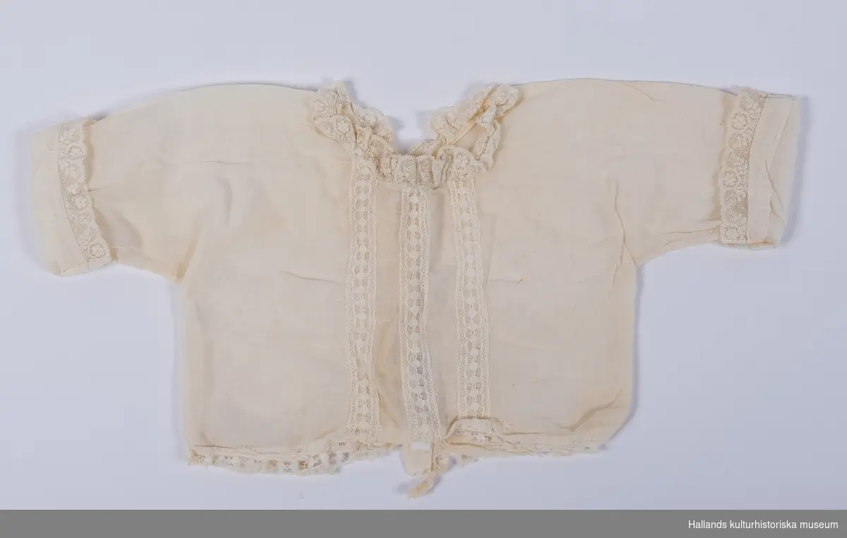 Babyskjorta av bomullsbatist. Spets runt ärmlinning, halsringning och tre spetsbårder framtill.