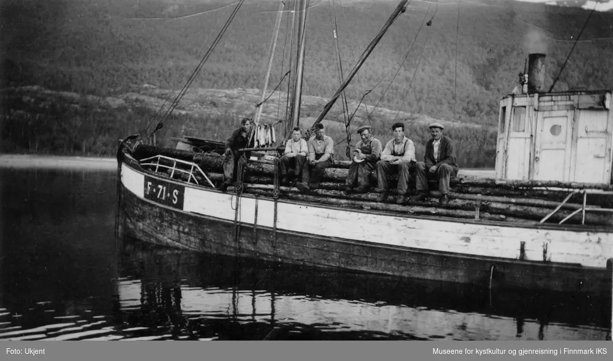 Seks personer sitter på tømmerlasset om bord på skøyta Ruth. Skøyta tilhørte Ragnvald Nicolai Normann fra Melkøya og de tok en fraktetur til Kvænangen for å hente trevirke til fiskehjeller. Båten har påskriften "F-71-S". I bakgrunnen ser man høye, med trær bevokste  fjell.