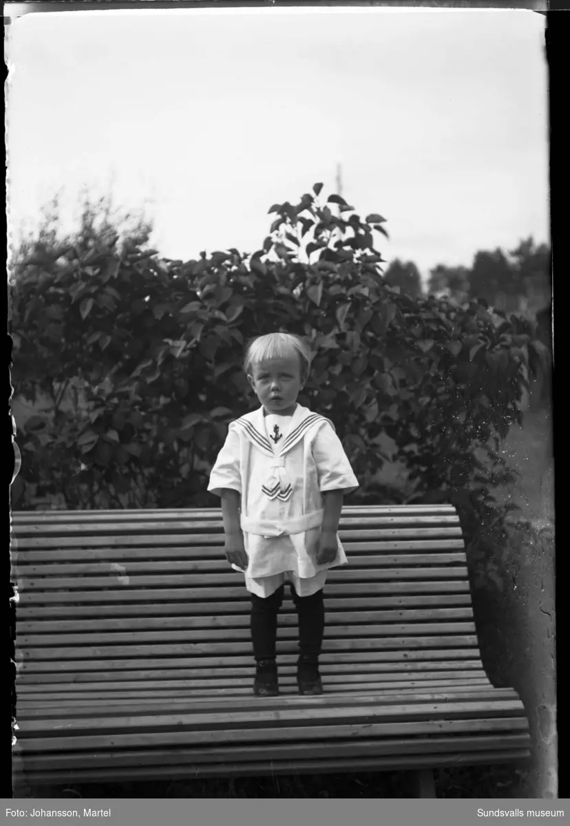 Porträttbild av en liten pojke i sjömanskostym, han står på en soffa ute i trädgården.