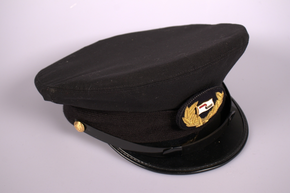 Svart uniformslue med skjerm. og  luemerke med rederiflagget til Det Nordenfjeldske Dampskibsselskab