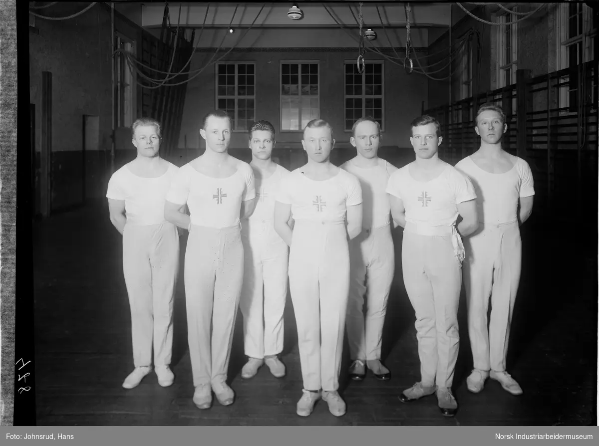 Mannlige turnere fra Snøgg, pokallaget fra 1929. Syv menn oppstilt i gymsal med turnbekledning. På brystet er emblemet for Norges Turnforbund. I bakgrunnen ribbevegger langs veggen og ringer for turn.