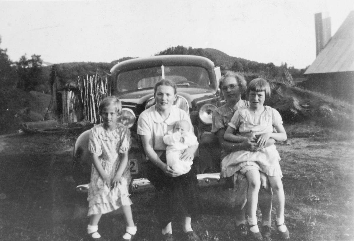 Fire jenter med en baby, fotografert på et gårdstun foran en bil.