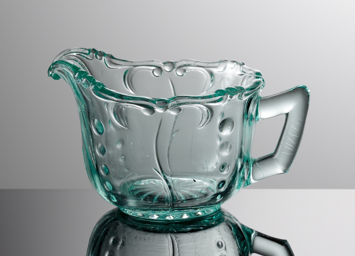 Pressad gräddkanna i ljusgrönt glas. Motiv av stiliserade växter i relief.