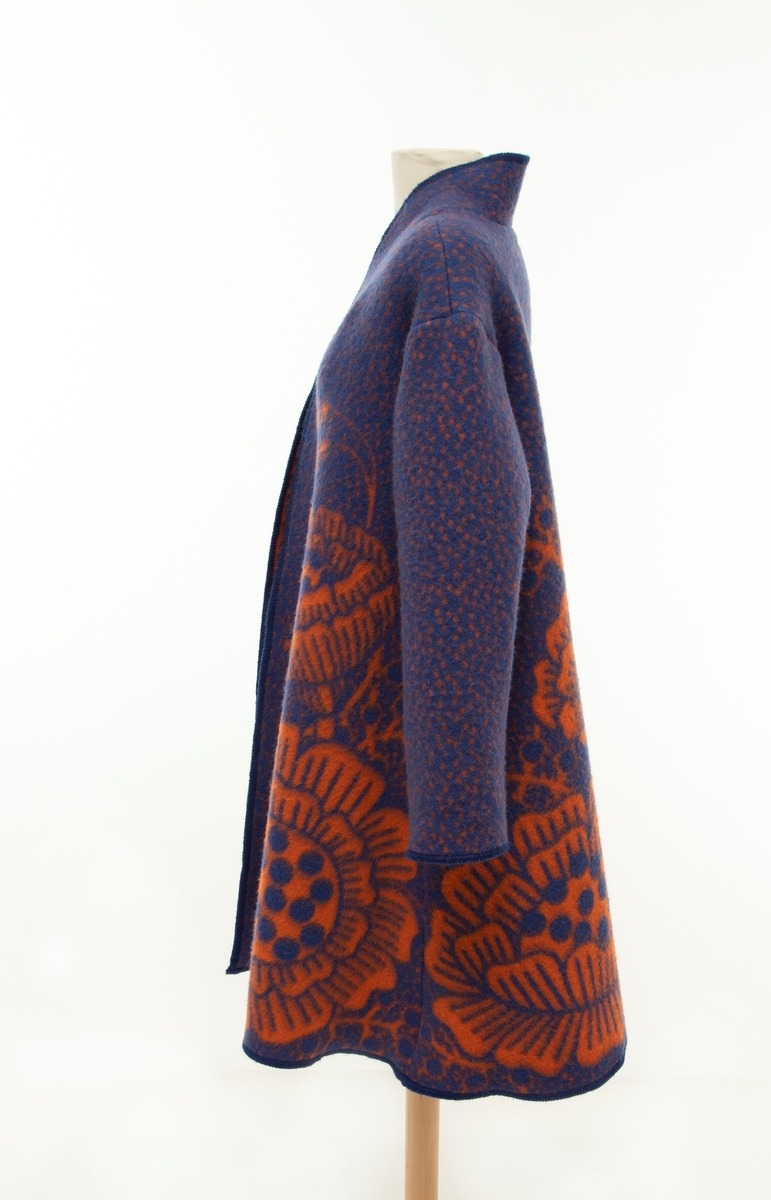 A-formet kåpe i blåfarget ull, dekorert med store orange blomster. Den har ståkrage, lav skuldersøm og sidelommer.