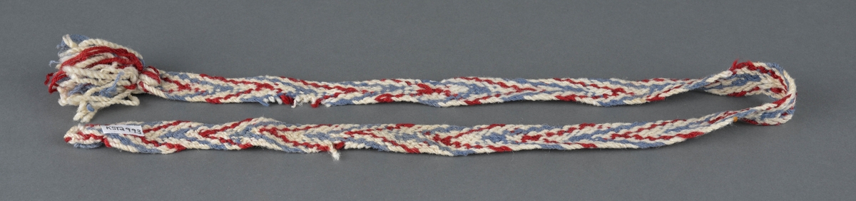 Strømpeband, hoseband fletta i rødt, blått og sauekvitt ullgarn.
Dusk i ene enden. Mangler den andre båndet.