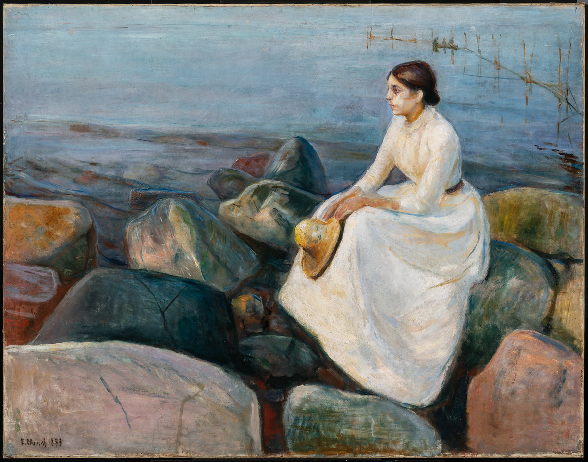 På en strandbredd mellom store steinblokker sitter en ung kvinne i hvit kjole med gul stråhatt i hendene, profil mot venstre. Blå sjø i bakgrunnen.