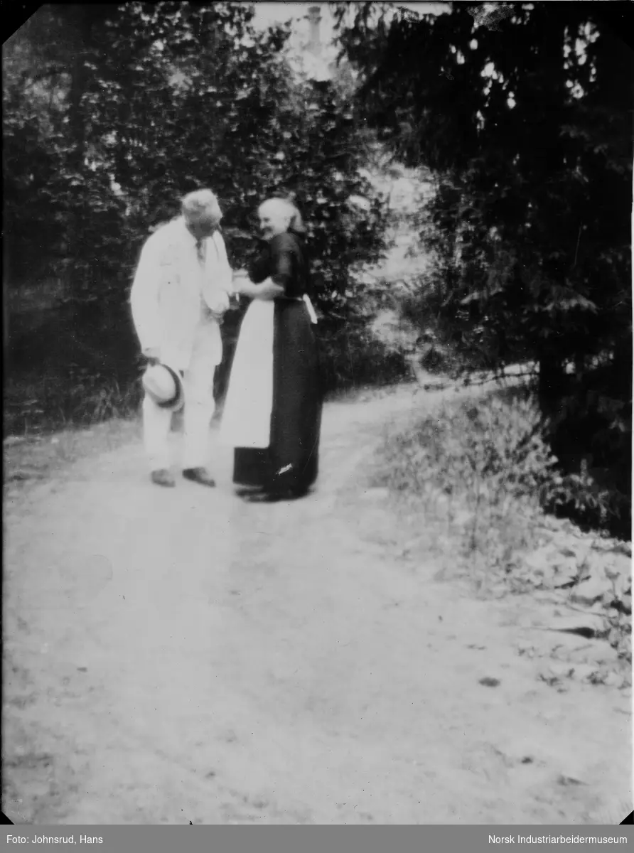 Mann og kvinne gående på vei. Mannen kledd i lys dress, kvinnen i mørk kjole med lyst forkle.