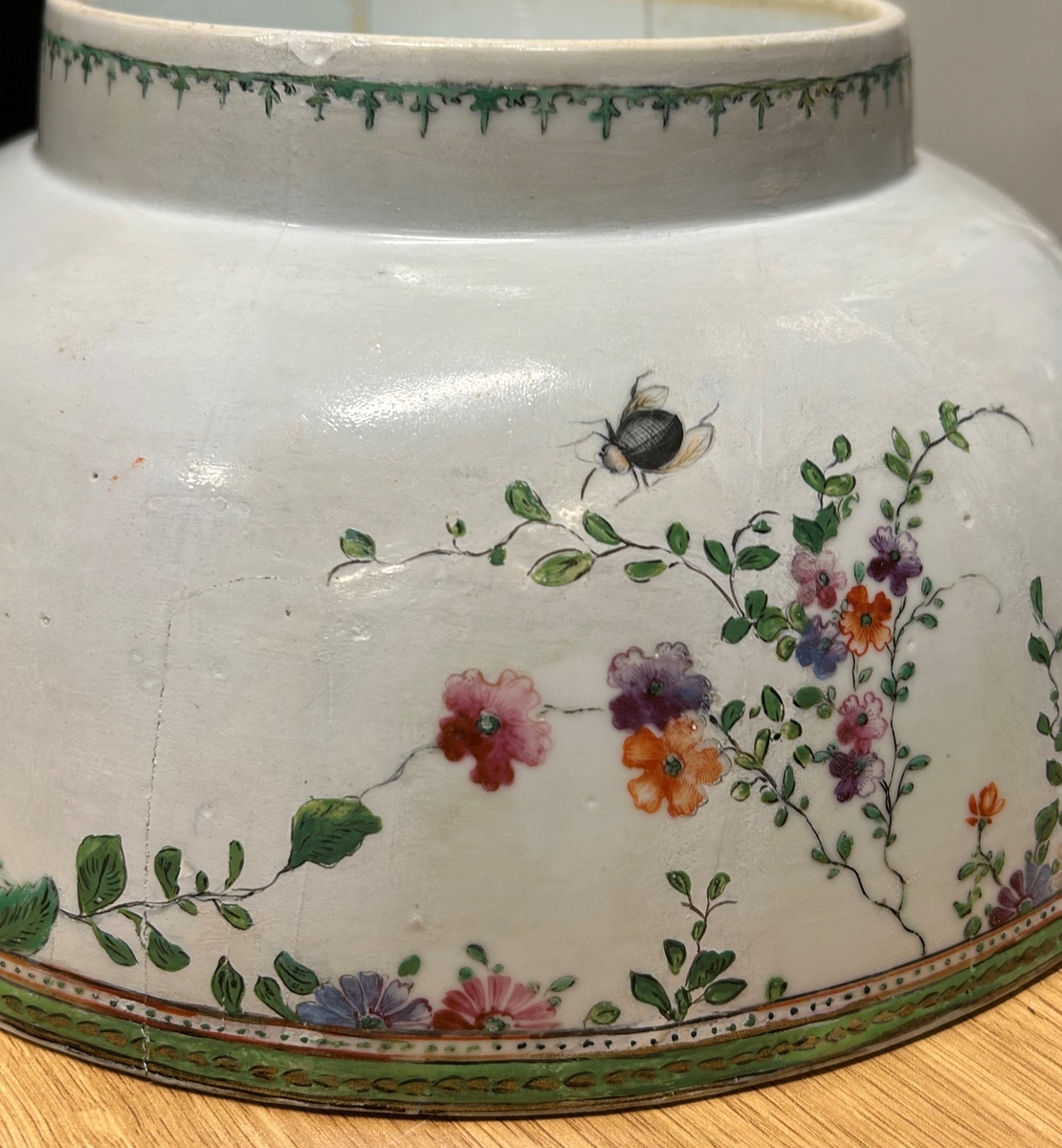 Vit cirkelformad skål av ostindiskt kompaniporslin med blommor i grönt, guld och rött, två fjärilar, en husfluga och en skalbagge. En markerad målad kant upptill utan och innanför på skålen. En blomma på botten av bålskålen. 1700-tal.