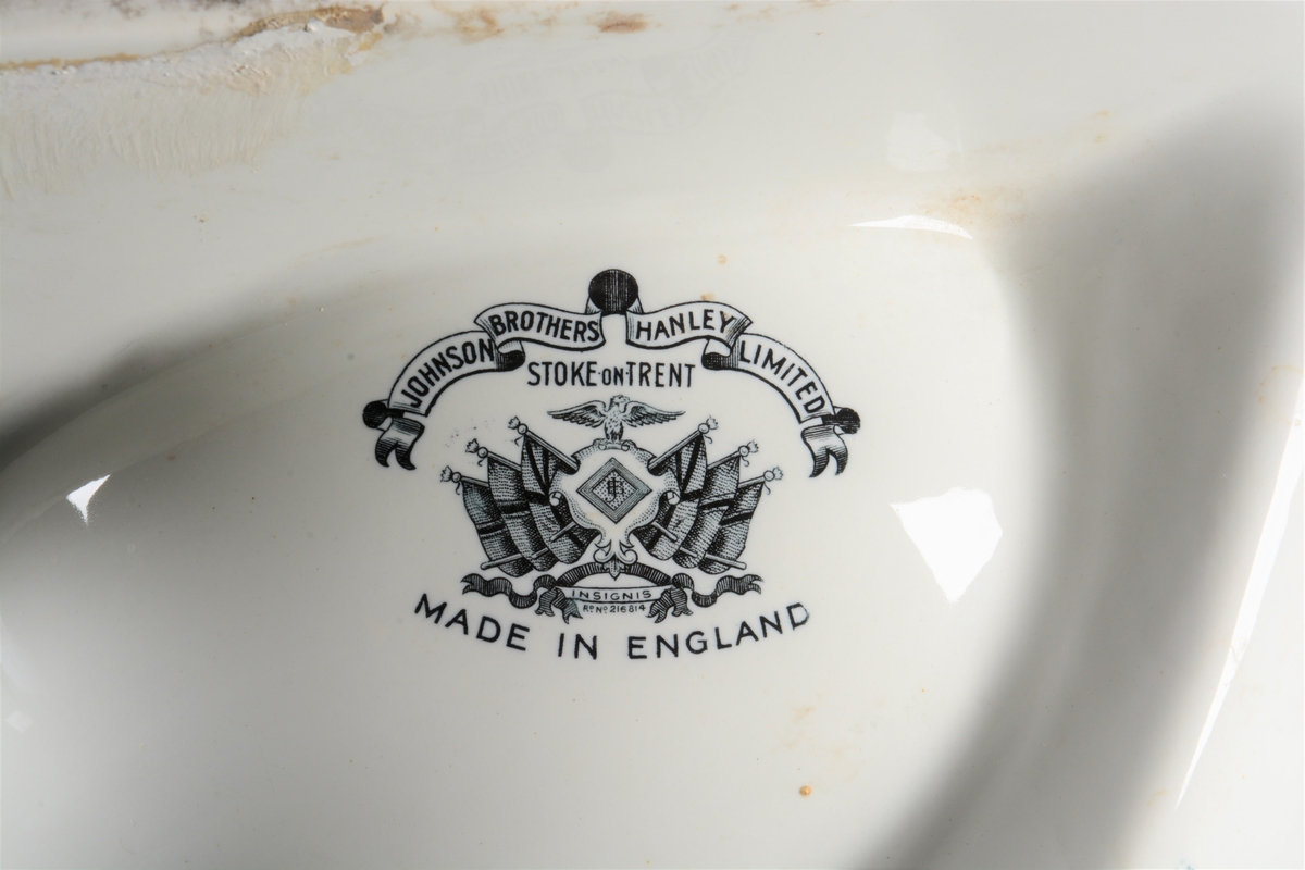 Hjørnevask av hvitt porselen. Den har rund skål og to rette sider med høy kant, slik at vasken passer i et hjørne. Vasken har sluk og rør av metall. På underiden av skålen er produksjonsstempel til firmaet "Johnson Brothers" med en ørn som holder et våpenskjold med initialene "JB", flankert av seks britiske flagg.