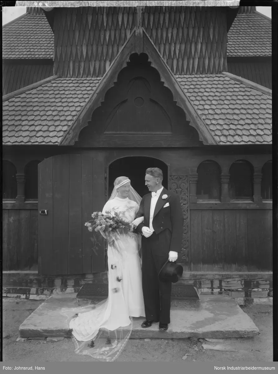 Brudepar foran Heddal Stavkirke. Kvinnen med brudebukett på armen, mannen med flosshatt i hånden. Bruden med slør og slep på kjolen. Knyttet til navnet Werge Olsen.