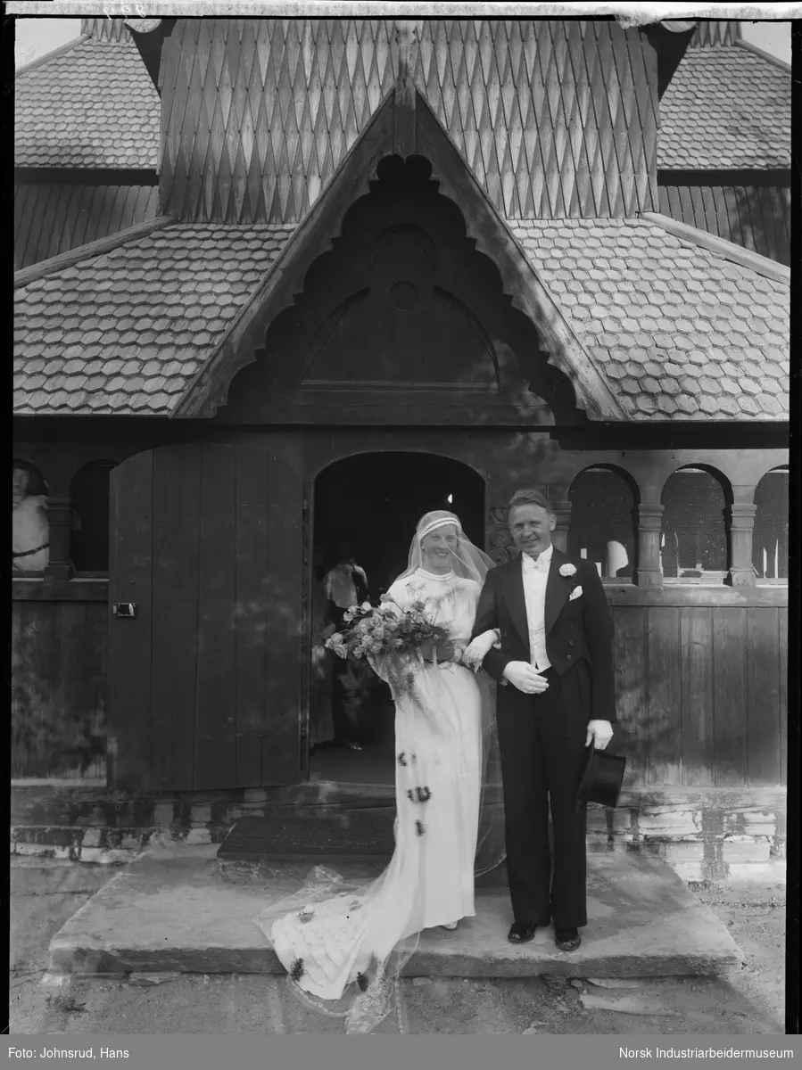 Brudepar foran Heddal Stavkirke. Kvinnen med brudebukett på armen, mannen med flosshatt i hånden. Bruden med slør og slep på kjolen. Knyttet til navnet Werge Olsen.