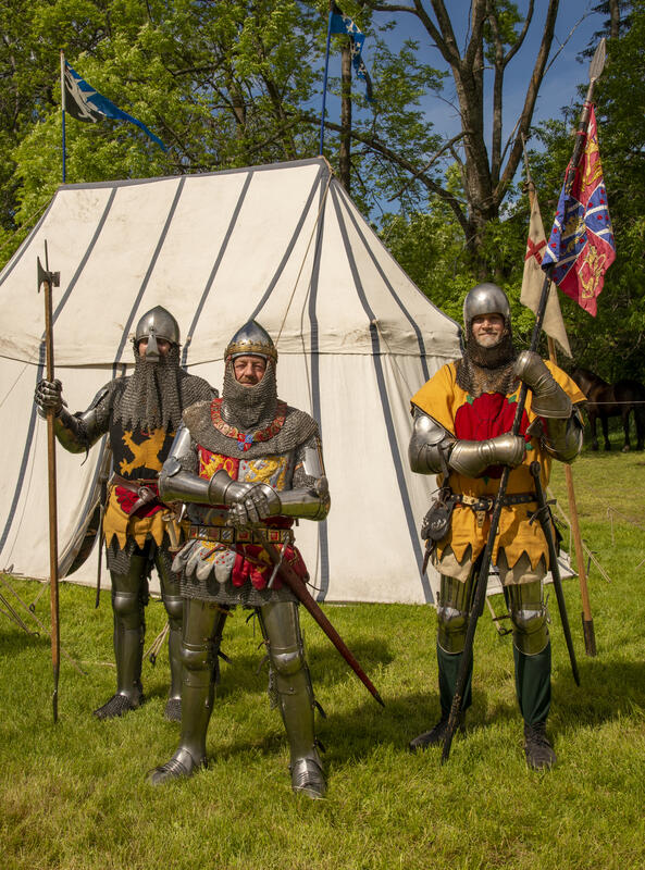 Tre riddere i platerustning med hjelmer og kjortler med våpenskjold i ulike farger brodert på kjortelen