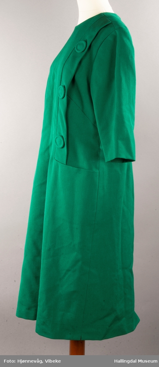 Grønn kjole med halvlange ermer. 3 store knapper på venstre side som er festet på en stolpe. Ei stor lomme under stolpen. Glidelås i ryggen. Rund hals.