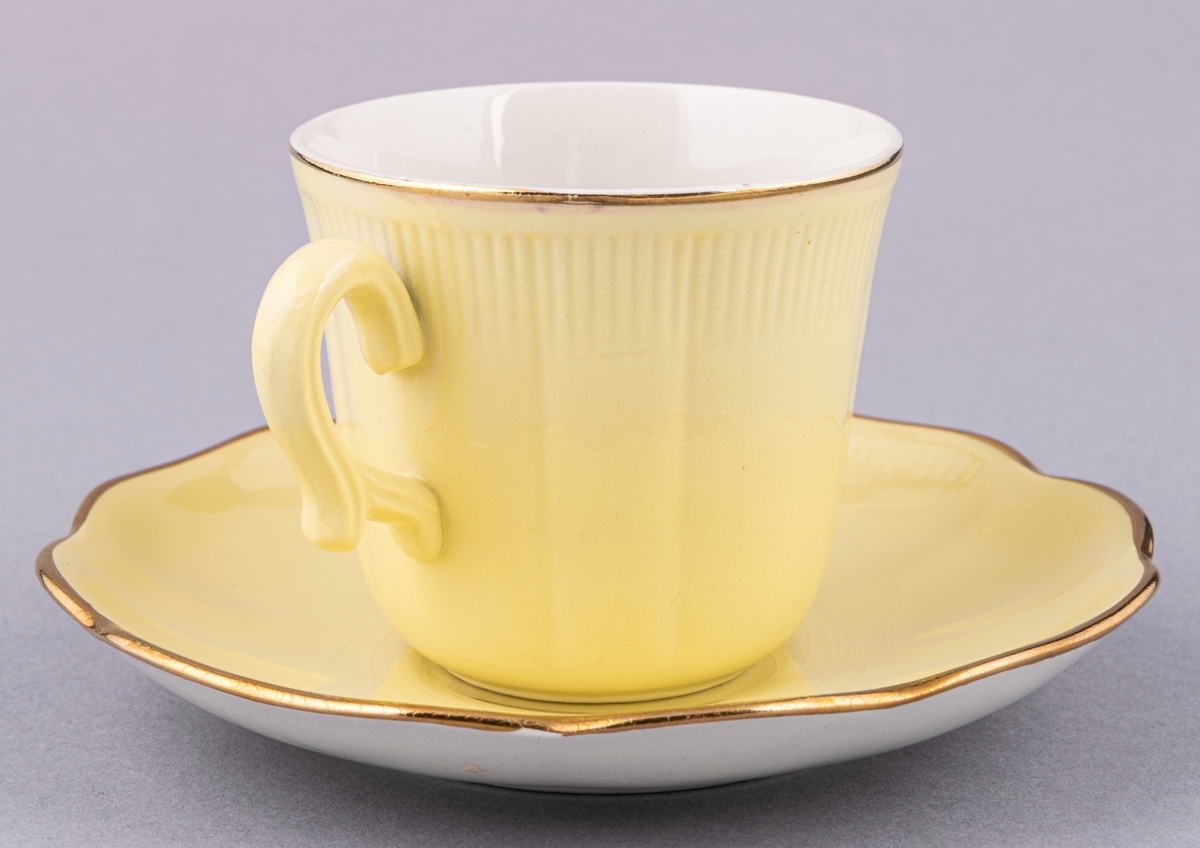 Kaffekopp med fat, flintgods. Modell AO, dekor Capri. Ljusgul glasyr på ovansidan, vit innuti, smal guldrand. Tillverkad vid Gefle Porslinsfabrik omkring 1949.