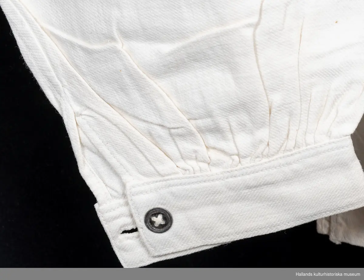 Skjorta av kypertvävd bomull, maskinvävd, maskinsytt. Lång ärm, fastsytt skjortbröst, slå med knäppning med två knappar baktill.