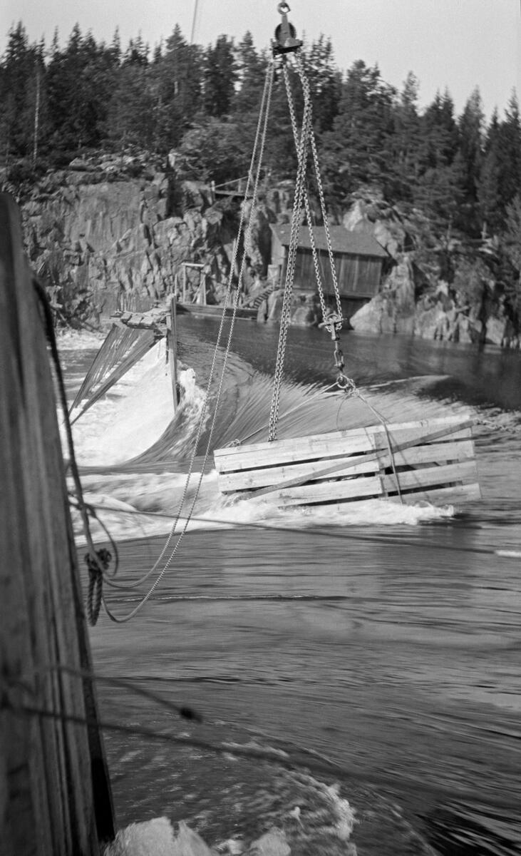 Bygging av såkalt fermettedam ved Mørkfoss i nedre del av Glomma høsten 1914.  Dette er en forholdsvis lett, men robust nåledamtype, der nålene holdes på plass av stålbukker som er hengslet til botnelementer på en måte som gjør det overkommelig å demontere dem om det skulle være påkrevet i reparasjonsøyemed.  Fotografiet er på tvers av vassdraget (fra Trøgstad-sida mot Skiptvet-sida), mot en åpning midt i elveløpet, hvor vannet fosset ut.  Over konstruksjonen ser vi ei taubane med løperkatt, som åpenbart ble brukt til å heise på plass damelementer.  Da dette fotografiet ble tatt var man i ferd med å senke steinkasser ned i den stride strømmen i det åpne midtløpet.  Disse skulle antakelig skulle tjene som tyngsel for konstruksjonen.  På motsatt side av denne åpningen i damkonstruksjonen, merd tyriangulætr stålbukker med ei gangbru på toppen, som fløterne kunne operere fra. På den bakenforliggende elvebredden ser vi et berglendt terreng med skog og ei lita bu med pulttakk, muligens ei redskapsbu. Dammen på fotografiet ble sannsynligvis demontert vinteren 1924, for fra påfølgende vår var det den da nye dammen ved Solbergfossen som regulerte Øyeren.

Den første fermettedammen ble konstruert av den franske ingeniøren Poirée i forbindelse med kanaliseringa av Mainz så tidlig som i 1831.  Dette anlegget ved Mørkfoss kan ha vært det første i sitt slag her til lands.  Denne dammen må ha virket som en terskel i vassdraget, i og med at den hevet vannspeilet oppover mot Øyeren, og dermed gjorde det enklere å få tømmeret fra Øyeren ned til Mørkfossen uten at det strandet på steinskjær og grusører, samtidig som det ble mulig å gå ned i dette området med små slepebåter. 

Fra gammelt av var Mørkfoss en vanskelig passasje for aktørene i tømmerfløtinga.  Vassdragsingeniøren Gunnar Sætren beskrev fossen og den effekten det avbildete damanlegget på stedet fikk slik (1904):

«Umiddelbart nedenfor Øieren er Mørkfossen.  Denne bestod oprindelig af tre fald, og da den var meget smal, havdes særdeles høie flomme i Øieren.  Dette forhold er nu betydelig forbedret, efterat der i aarene 1857 til 69 blev udført betydelige sprængningsarbeider til utvidelse af Mørkfossen.  Samtidig opførtes en reguleringsdam ovr Mørkfossen for at hæve lavvandet i Øieren af Hensyn til dampbaadsfarten og flødningen.  Ved disse arbeider er store flomme sænket betydelig ....»