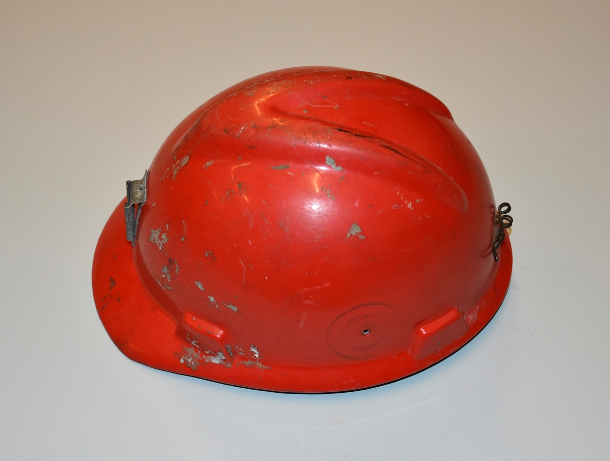 Hjelm i rød plast med fester i metall for lys foran og ledning til batteri bak. Innmaten er laget av plast og nylonreimer.