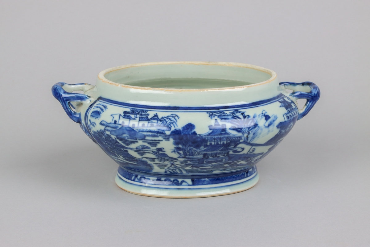 Oval, lobed bowl with angular handles, supported on a central foot. On the both sides decor of landscape scenes of pagodas, buildings, gardens and waters. Handles in shapes of bent tree branches. On the foot a  dark blue border with rectangular reserves filled with symbols of fortunes. The base of the bowl  with unglazed ring.
