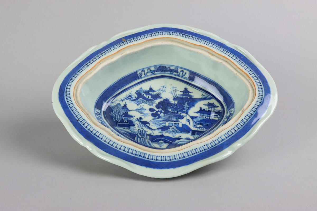 Oval broken form with a wide flat rim, decorated with a dark blue border with criss cross pattern. The inside of the dish slightly everted and undecorated. The well surrounded with a border of a criss cross pattern and rectangular reserves filled with symbols of good fortune. Centered in the well landscapes scenes with pagodas, buildings, waters, bridges and gardens. The outside of the dish is decorated with rose branches. All decor in blue underglaze. The base of the dish without glazing.