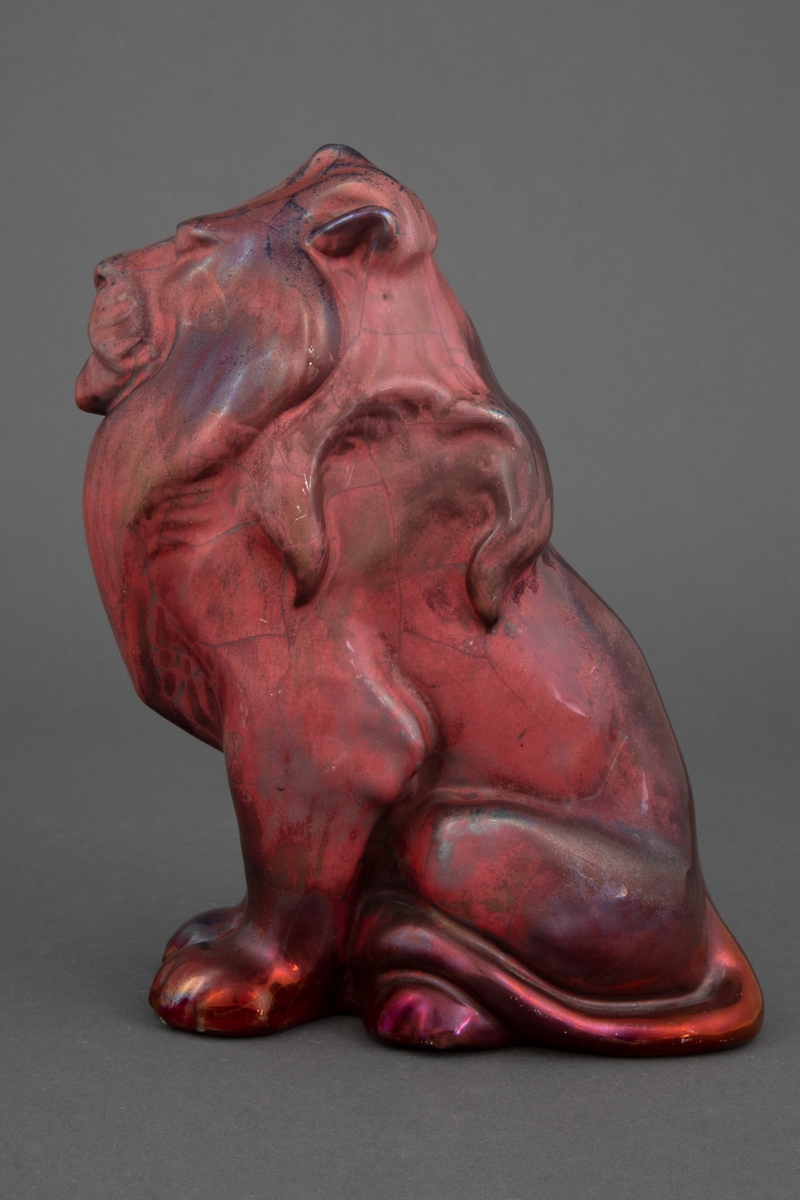 Keramikkfigur som viser en sittende løve. Løven er i lett, lysegrått gods. Figuren er glasert med mørkerød blank glasur og metall-luster.