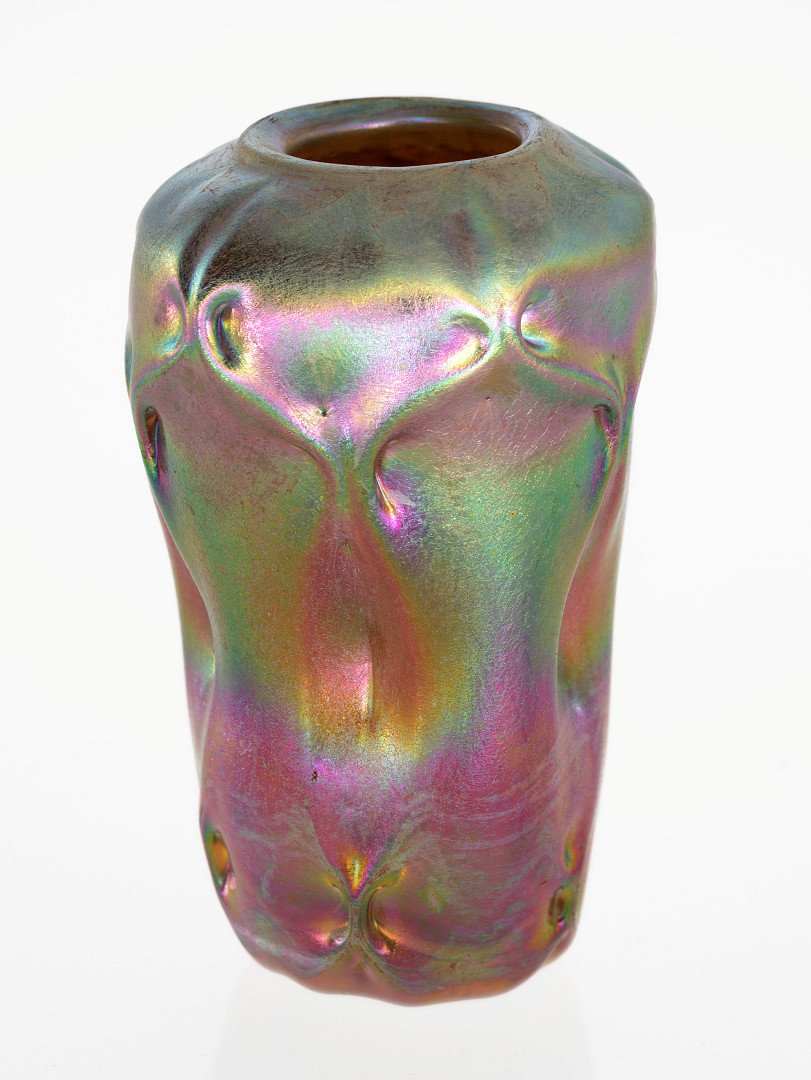 Konisk vase i irisert glass med organisk utforming. Vertikalt svugne riller på korpus. Den skimrende overflaten veksler mellom grønne, gule og blålilla partier. Sirkulær svakt hevet munningsrand.