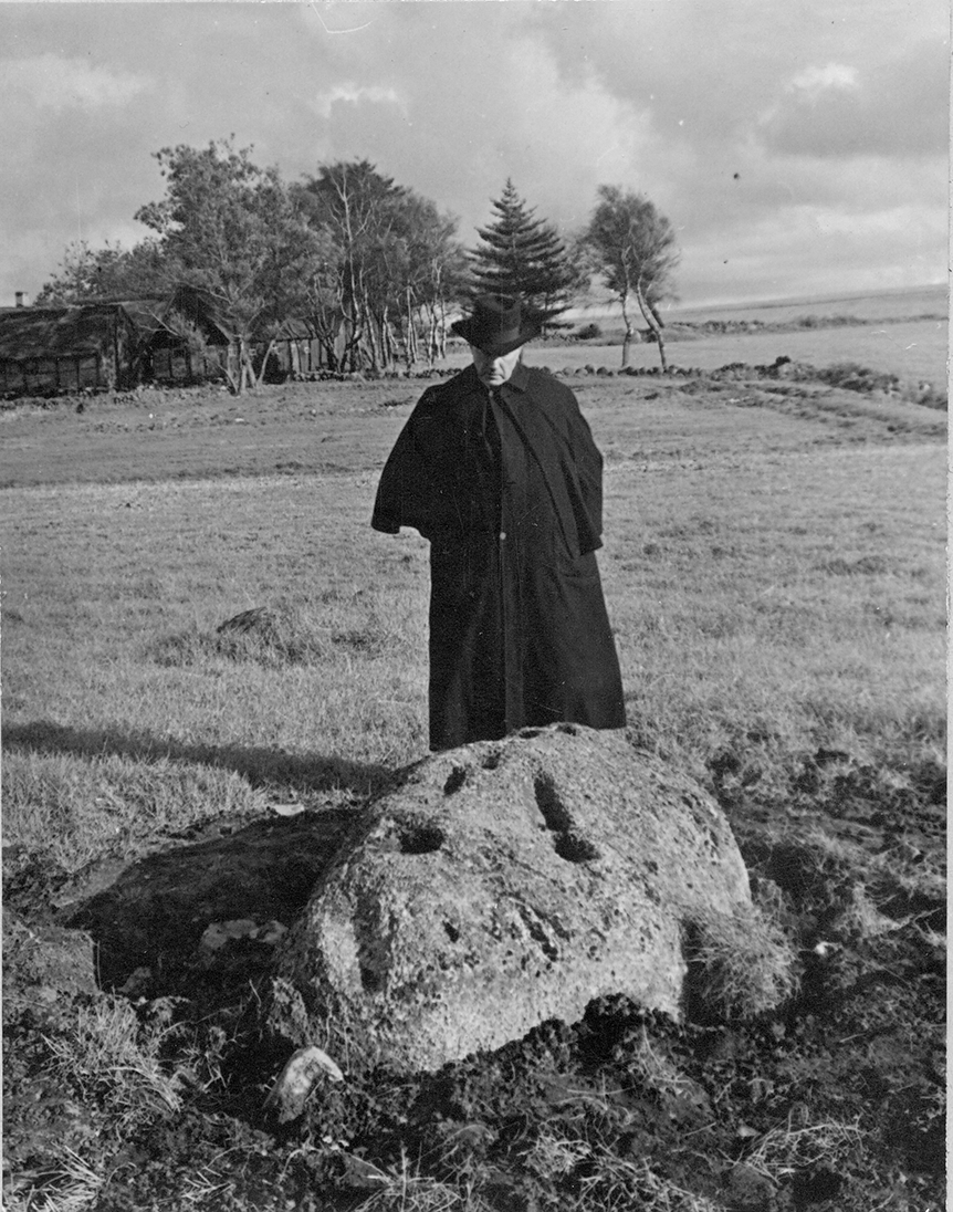 Sten med hällristning och skålgropar i Tjuvahålan Hasslöv. Vid stenen står en man klädd i mörk slängkappa och hatt. I bakgrunden ligger en korsvirkesgård i en dunge varifrån stengärdsgårdar löper ut över ägorna.