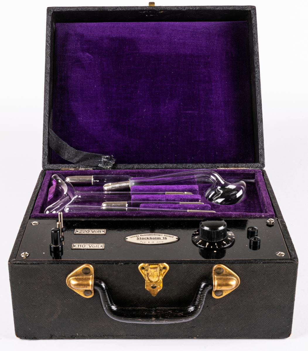 Elbehandlingsapparat i nio delar.
Svart väska med insidan klädd i lila sammet, svart bakelithandtag samt 5 tillbehör i glas o metall.